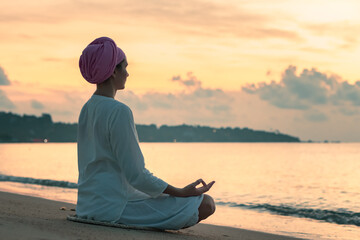 A woman in white clothes and a turban sits by the sea on a sandy beach at dawn on a summer morning...
