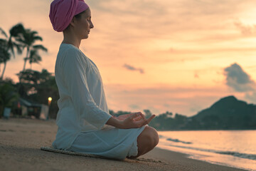 A woman in white clothes and a turban sits by the sea on a sandy beach at dawn on a summer morning and doing yoga exercises . Colorful sky on the background.