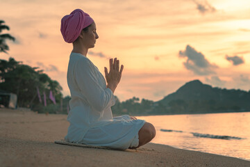 A woman in white clothes and a turban sits by the sea on a sandy beach at dawn on a summer morning and doing yoga exercises . Colorful sky on the background.