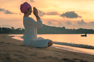 A woman in white clothes and a turban sits by the sea on a sandy beach at dawn on a summer morning and doing yoga exercises . Colorful sky on the background.