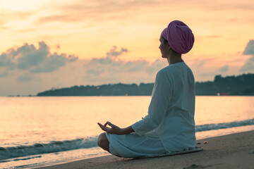 A woman in white clothes and a turban sits by the sea on a sandy beach at dawn on a summer morning and doing yoga exercises . Colorful sky on the background.