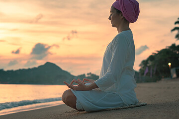 A woman in white clothes and a turban sits by the sea on a sandy beach at dawn on a summer morning and doing yoga exercises . Colorful sky on the background.