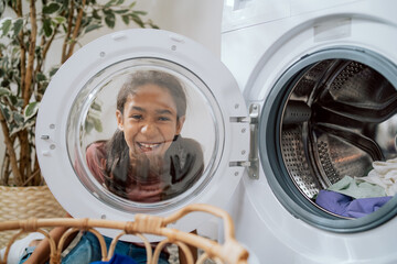 A cute smiling little girl with brown hair and dark eyes looks through the round glass door of the washing machine, watches her mother sorting laundry in the bathroom