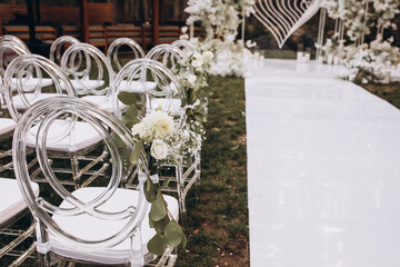 Wedding. Ceremony. There are white transparent designer chairs for guests on a green meadow in the forest. Chairs decorated with white flowers