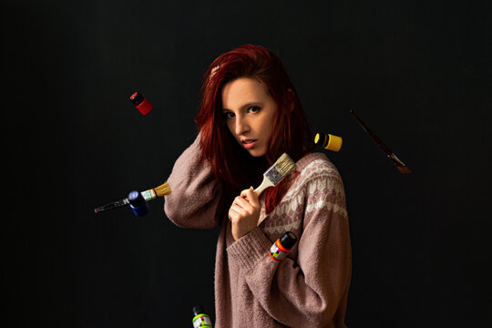 The artist at the moment of creativity: a beautiful red-haired skinny girl in a knitted sweater holds a brush in her hand, tubes of paint and brushes are flying around her. Metaphor of the idea