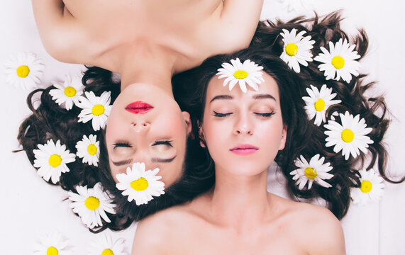 Two trendy topless twin sisters cheerful summer hipster teenage girls in a spa salon lie naked face to face on their backs covered in chamomile flowers. The concept of skin care, health and motherhood