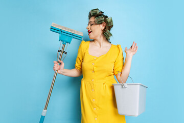 Comic portrait of young emotional girl, housewife with curlers on her head isolated on blue studio...