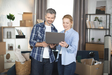 Modern Caucasian adult couple smiling and enjoying relocation at new flat, standing with laptop computer and making online shopping. Moving to modern apartment. Cardboxes around.