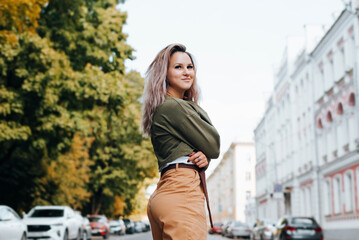 Naklejka premium Pretty hipster in youthful casual clothes with cute smile standing on street and looking away. Half-length portrait of young caucasian slim woman posing in city at daytime outdoors. Summer lifestyle