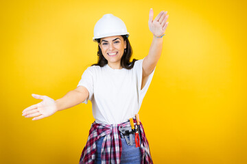Young caucasian woman wearing hardhat and builder clothes over isolated yellow background looking at the camera smiling with open arms for hug