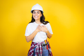 Young caucasian woman wearing hardhat and builder clothes over isolated yellow background smiling with her hands on her chest and grateful gesture on her face.