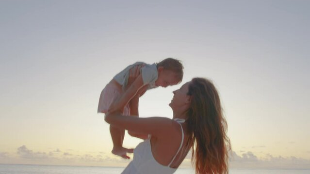 Authentic close up shot of young mother is keeping on her arms and playing with a newborn baby on a seaside beach at sunset during holiday vacation