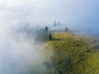 Fog envelops the mountain forest. The rays of the rising sun break through the fog. Aerial drone view.