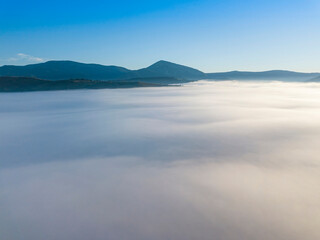 Naklejka premium Flight over fog in Ukrainian Carpathians in summer. Mountains on the horizon. A thick layer of fog covers the mountains with a continuous carpet. Aerial drone view.