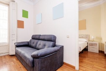 Living room with a balcony with white wooden shutters, a black leather two-seater sofa next to an...