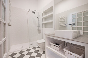 Toilet with built-in washbasin cabinet with white porcelain washbasin, wicker baskets, mirror without frame and shower cabin, wooden door with opaque glass and checkerboard stoneware floors