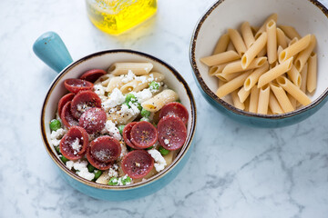 Turquoise bowls with penne pasta, crispy salami, ricotta cheese and green peas, elevated view on a light-grey marble background, studio shot
