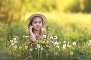Cute little girl in a straw hat has sits on a summer field with dandelions. Child having activity fun outside. Concept of a healthy child without allergies. High quality photo. High quality photo