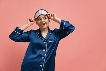 Half-length portrait of young cheerful woman in silk pajamas wearing sleeping mask, posing isoated...