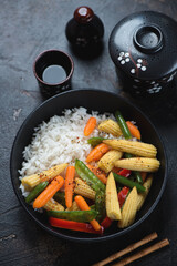 Black bowl with vegetable stir-fry and rice, vertical shot on a dark-brown stone background, elevated view