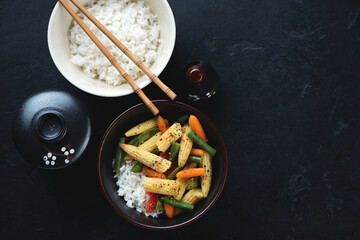 Bowls with stir-fried vegetables and white rice, above view on a black stone background, horizontal shot with copy space