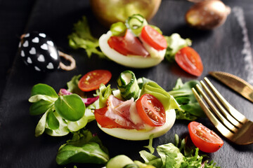 Easter breakfast with egg halves with prosciutto ham and vegetables on black board closeup