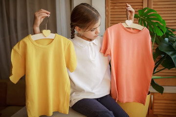 Pretty girl chooses T-shirt among clothes at home, she holds light colored yellow and pink plain...