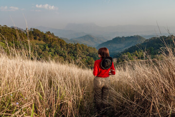 A woman in a red long-sleeve with a white hat walks on golden dry grasslands at Hadubi, Chiang Mai,...
