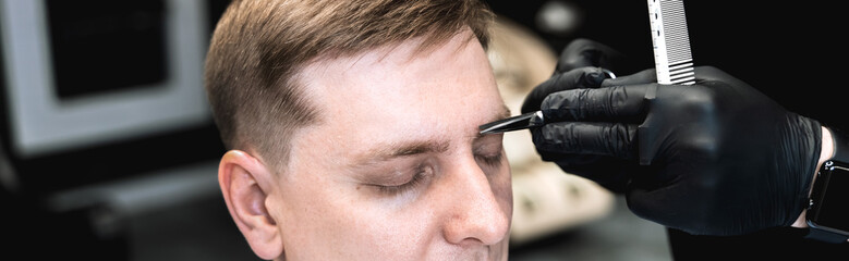 Obraz premium Visit to barbershop.Eyebrow trimming with scissors.Stylish man makes fashionable haircut.Barber,hairdresser,stylist with comb,dryer.Combing hair.Client,customer sits in salon covered with cape