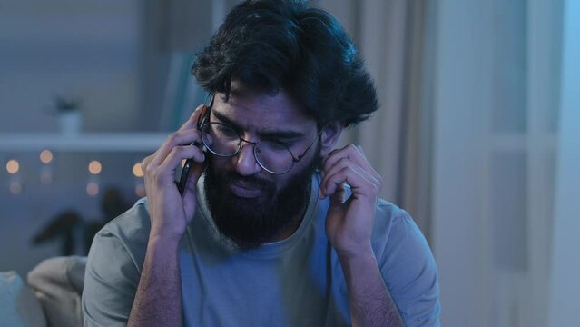 Arabic Indian bearded man with glasses talking on phone at house party sitting on sofa neon light hard to hear conversation loud music noisy birthday celebration answering phone call trying listening