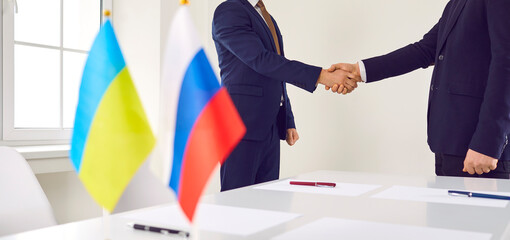 Moscow and Kyiv representatives regulate conflict, reach peace settlement and shake hands over negotiation table with two national flags. Russia ceases shelling, stops war, ends Ukraine state invasion