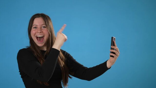 Surprised young woman 20s posing isolated on blue background studio. Using mobile cell phone typing sms message pointing index finger to copy space say wow