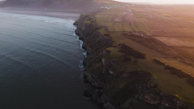 AERIAL: Dolly along cliff face coastline during hazy sunrise, Rhossili Gower, 4k Drone