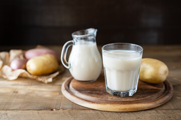 Vegan potato milk in glass and milk jar on cutting board with raw potatoes aside. Wooden background. Rustic style