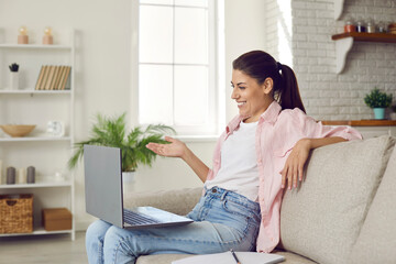 Woman using laptop computer for online communication or distance learning. Happy positive young girl sitting on sofa at home, talking to friend, tutor or colleague on video call, gesturing and smiling