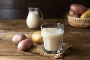 Vegan potato milk in glass and milk jar with raw potatoes on the wooden background in rustic style