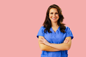 Portrait of smiling female doctor or nurse in blue uniform with arms crossed on pink studio...