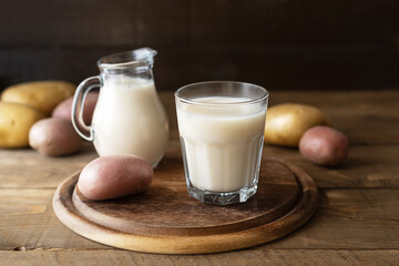 Vegan potato milk in glass and milk jar on cutting board with raw potatoes aside. Wooden background. Rustic style