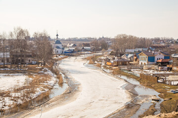 A crooked, frozen river stretching into the distance, wooden houses on the shore. Spring, snow melts, puddles and dry grass all around. Day, cloudy weather, soft warm light.
