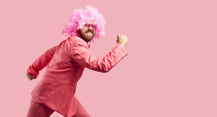 Happy goofy clown having fun in the studio. Funny handsome young man wearing pink wig, trendy suit and cool sunglasses running forward very fast isolated on solid pink color background