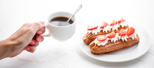 Woman's hand holding a cup of coffee over Eclairs with cream and strawberries on a white table - Powered by Adobe