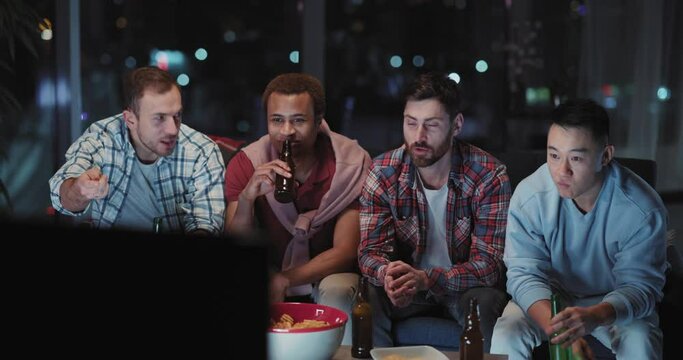 Diverse group of crazy football fans watching TV at night. Multi-race friendly young men drinking beer sharing stories communicating. Friendship concept.