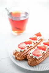 Eclairs with cream and strawberries and a cup of tea on a white plate. 