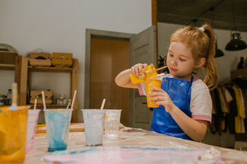 Child girl sitting in art school at the table with paints mixing yellow color