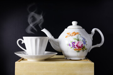 Teapot, cup with steam on wooden table on dark background, white porcelain service