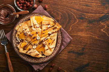 Crepes suzette with oranges and chocolate. Thin crepes with chocolate spread, hazelnuts and orange slices fruit in plate for breakfast on old dark wooden rustic table background. Copy space. Top view