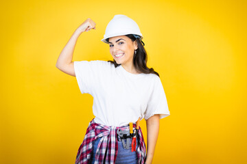 Young caucasian woman wearing hardhat and builder clothes over isolated yellow background showing arms muscles smiling proud