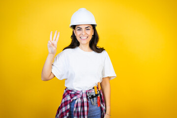 Young caucasian woman wearing hardhat and builder clothes over isolated yellow background showing and pointing up with fingers number three while smiling confident and happy
