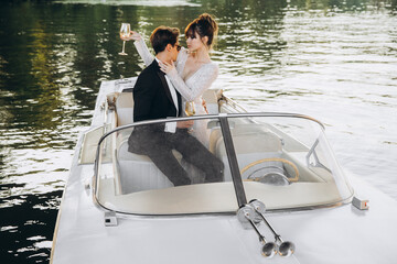Couple rides a yacht on a sunny day. Stylish groom in a black suit and sunglasses hugs and kisses a...