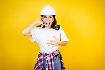 Young caucasian woman wearing hardhat and builder clothes over isolated yellow background confused doing phone gesture with hand and fingers like talking on the telephone